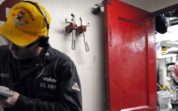 Two U.S. Navy personnel are photographed aboard what appears to be the USS Fitzgerald (DDG-62), a naval vessel. One sailor in a black uniform and yellow USS Fitzgerald cap is writing on a clipboard, while a second person in the background wears full protective gear including a blue suit, red helmet, and white gloves. The setting appears to be an interior compartment or workspace on the ship.