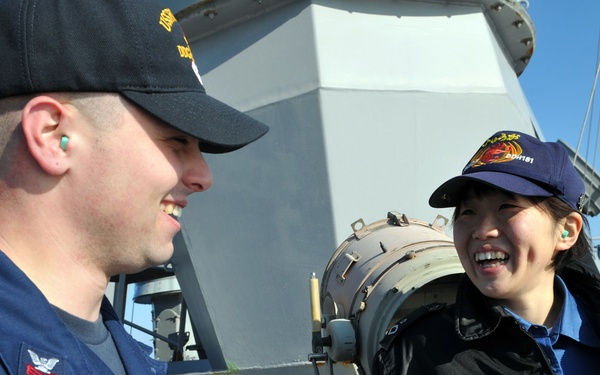 Two naval personnel are engaged in a friendly conversation on the deck of a warship. The male sailor on the left wears a US Navy uniform with the name tag 'WHALEN' and a dark cap, while the female sailor on the right wears a navy blue cap with what appears to be a Japanese Maritime Self-Defense Force insignia. Both are smiling and wearing ear protection (earplugs), suggesting they are in or near a high-noise environment.