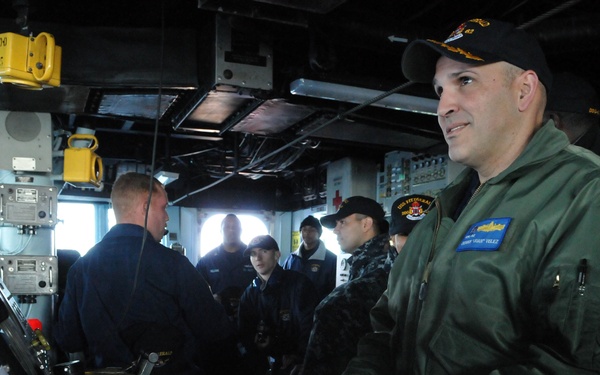 The image shows military personnel aboard what appears to be a naval vessel's bridge or combat information center. A prominent figure in the foreground wears a green flight jacket with a name tag and a black military cap with insignia. Several other uniformed personnel are visible in the background engaged in various duties.