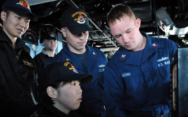 The image shows U.S. Navy personnel and what appear to be Japanese Maritime Self-Defense Force members aboard a U.S. Navy vessel, likely the USS Fitzgerald (DDG-62) based on visible cap insignia. A U.S. Navy sailor with the name tag 'HOAR' is demonstrating or instructing others on the operation of ship controls or equipment. The scene appears to be a training or familiarization exchange between allied naval forces.