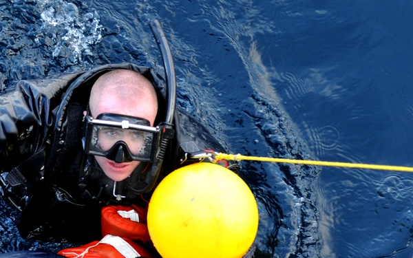 A rescue diver in a black wetsuit and diving mask is in open ocean water, holding onto a yellow buoy attached to a yellow tow line. The diver appears to be supporting a second person wearing an orange immersion/survival suit, suggesting a water rescue operation in progress. The scene depicts what appears to be a maritime search and rescue exercise or actual rescue operation.