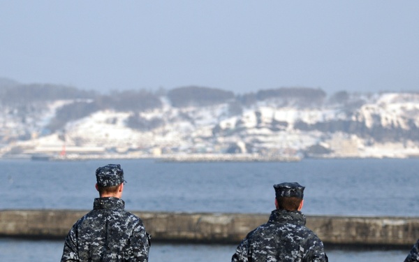 Four U.S. Navy sailors in Navy Working Uniform (NWU) Type I digital camouflage stand at attention on the deck of a naval vessel, facing toward a snow-covered coastline across a body of water. They appear to be in a formal 'man the rails' or observation posture, with hands clasped behind their backs. The setting suggests a cold-weather port environment, likely in the Pacific region based on the terrain.