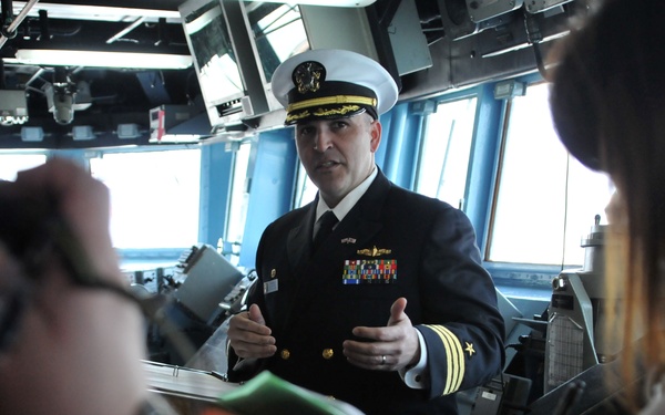 A U.S. Navy officer in dress uniform is speaking to a group of civilians or media personnel on what appears to be the bridge of a naval vessel. The officer is wearing a white-topped combination cover and a dark service dress uniform adorned with multiple ribbons and gold insignia. The setting suggests a briefing, tour, or press engagement aboard a warship.