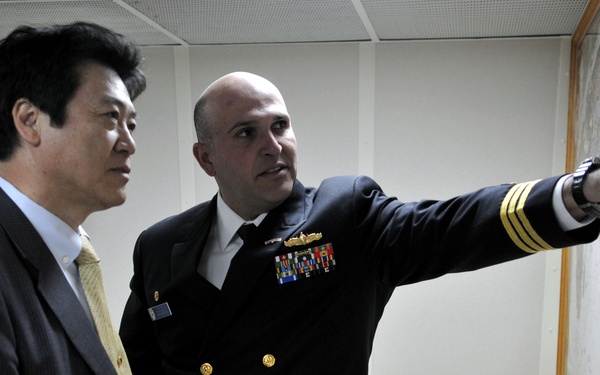 Two men are standing together examining a large framed map mounted on a wall. One man is wearing a dark business suit with a light tie, while the other is dressed in a dark naval officer's uniform with gold buttons, rank stripes, and multiple service ribbons. The naval officer is pointing at a specific location on the map.