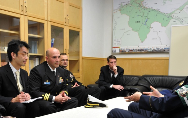 A formal military meeting is taking place in an office setting, with several uniformed and civilian-dressed individuals seated on black leather sofas around a white-clothed table. The group appears to include U.S. Navy officers and Japanese military or government officials engaged in a briefing or diplomatic discussion. Maps and charts are visible on the walls in the background.