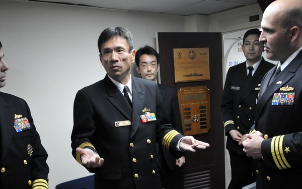 A group of naval officers in dress uniforms are gathered in what appears to be a ship's wardroom or conference room. The central figure, appearing to be of Asian descent, is gesturing with open hands as if explaining something to the officers flanking him. The setting suggests a formal military meeting or exchange visit between naval personnel.