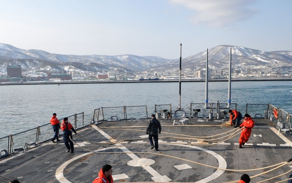 A naval vessel's flight deck is shown with numerous crew members wearing orange immersion suits and navy blue uniforms handling thick mooring lines, suggesting a docking or undocking operation. The ship appears to be in a harbor with a snow-covered mountainous coastal city visible in the background. A tugboat is visible in the water nearby, consistent with a port arrival or departure maneuver.