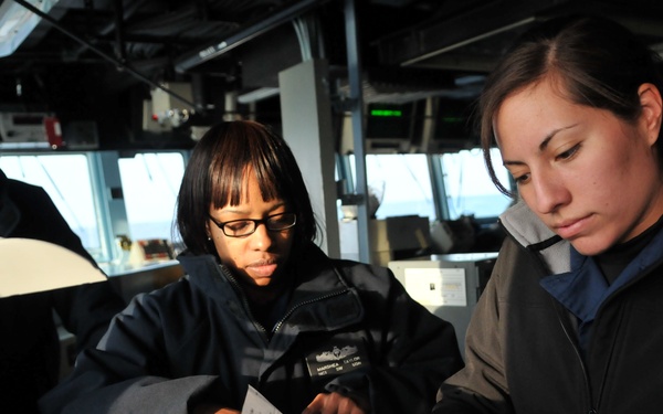 Three U.S. Navy personnel are on the bridge of the USS Fitzgerald (DDG-62), a guided-missile destroyer. Two female sailors in the foreground are reviewing documents and paperwork at a navigation console, while a male sailor in a Navy cap observes in the background. The setting appears to be an active ship bridge with navigation equipment and displays visible.