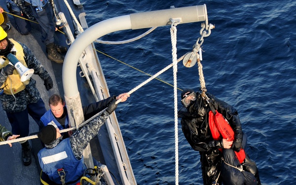 A naval rescue or training operation is underway on the deck of a military vessel, with a diver in a black wetsuit being hoisted by a crane/davit system over open ocean water. Several crew members in military uniforms and safety gear are managing ropes and equipment on the ship's deck. The diver is equipped with orange flotation devices, fins, and a diving mask, suggesting a search-and-rescue or man-overboard drill.