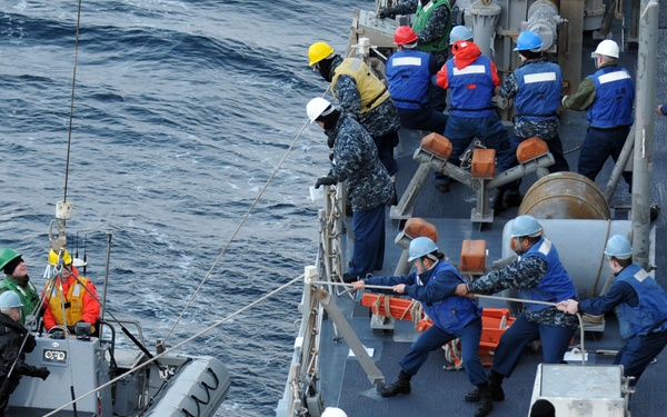 A naval vessel crew is conducting a small rigid inflatable boat (RIB) recovery or deployment operation at sea. Multiple personnel in military uniforms and safety gear are working together on the ship's deck, managing lines and rigging to hoist or lower the gray RIB alongside the vessel. The operation involves coordinated teamwork between crew members on deck and those aboard the small boat.