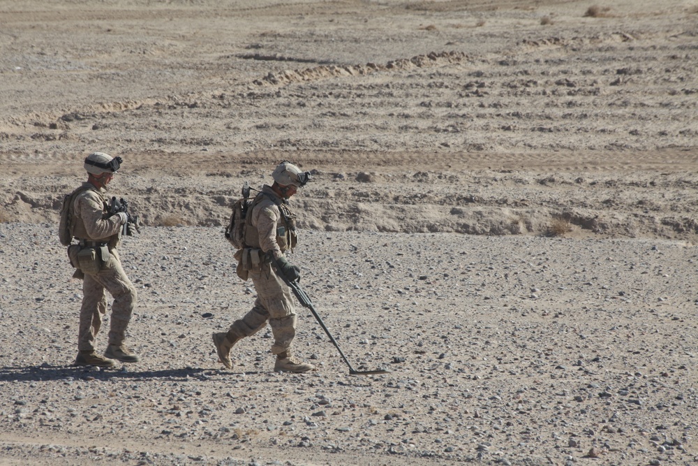 Two military personnel in desert camouflage uniforms are conducting what appears to be an IED or mine detection sweep across an arid, rocky terrain. The forward soldier is operating a handheld metal detector while both carry tactical gear and equipment. The environment is a flat, open desert landscape with sparse vegetation.