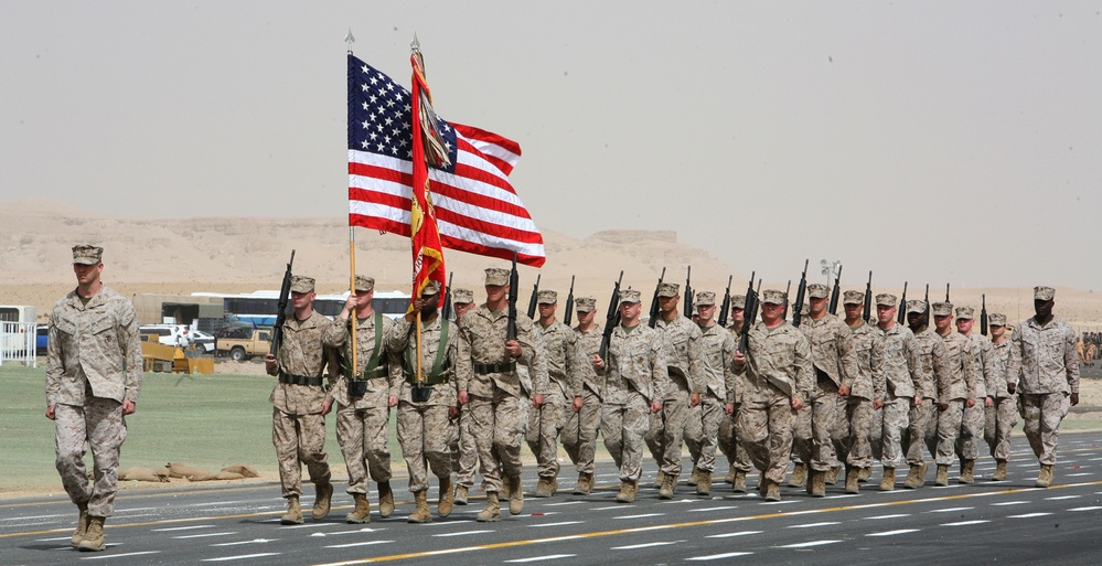 A formation of U.S. Marine Corps personnel marches in a formal parade or ceremony in what appears to be a desert environment. The troops are carrying the American flag and a Marine Corps unit guidon, with rifles at shoulder arms. The arid, sandy landscape and hazy sky suggest a Middle Eastern or desert deployment location.