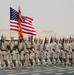A formation of U.S. Marine Corps personnel marches in a formal parade or ceremony in what appears to be a desert environment. The troops are carrying the American flag and a Marine Corps unit guidon, with rifles at shoulder arms. The arid, sandy landscape and hazy sky suggest a Middle Eastern or desert deployment location.