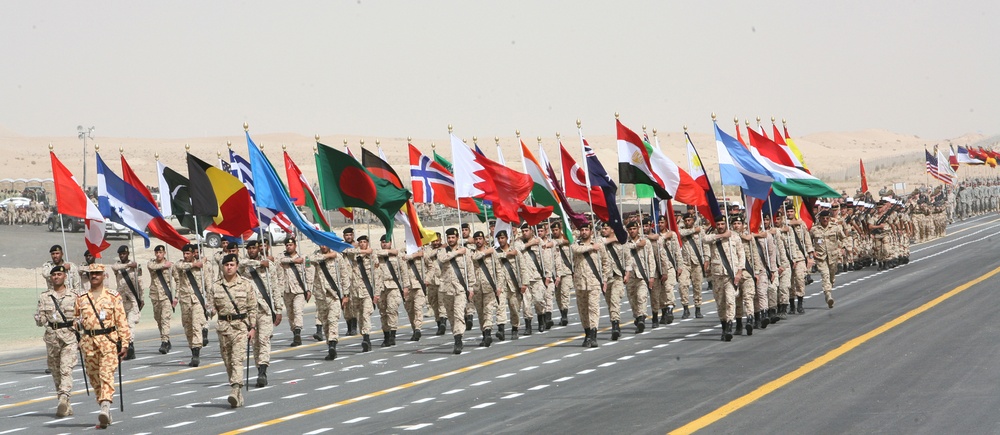 A large multinational military parade is taking place on a wide paved road in an arid desert environment. Soldiers dressed in tan/desert camouflage uniforms march in formation while carrying numerous national flags from various countries. The scale of the formation and diversity of flags suggests a significant joint military exercise or ceremonial event.