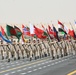 A large multinational military parade is taking place on a wide paved road in an arid desert environment. Soldiers dressed in tan/desert camouflage uniforms march in formation while carrying numerous national flags from various countries. The scale of the formation and diversity of flags suggests a significant joint military exercise or ceremonial event.