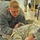 432nd Civil Affairs Battalion prepares for deployment to Afghanistan A soldier in U.S. Army ACU (Army Combat Uniform) and gray fleece jacket is kneeling over another soldier lying on the floor, appearing to perform a medical training exercise such as casualty assessment or CPR practice. The setting appears to be an indoor facility, likely a training room, with additional military personnel visible in the background. This appears to be a combat lifesaver or first aid training scenario.