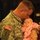 432nd CA BN prepares for deployment to Afghanistan A U.S. Army soldier in ACU (Army Combat Uniform) is tenderly kissing a newborn or very young infant dressed in pink clothing. The soldier's uniform displays a U.S. flag patch, a 1st Infantry Division ('Big Red One') patch, and a name tape partially reading 'LLER' (likely 'MILLER' or similar). The soldier also wears a wedding ring, suggesting this is likely a family reunion or deployment-related moment.