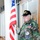 432nd CA BN prepares for deployment to Afghanistan An elderly man, identified by his patches and cap as a Vietnam Veteran, stands holding an American flag in what appears to be an indoor corridor or lobby. He is wearing a camouflage military-style jacket adorned with multiple patches and pins, including a memorial patch reading 'In Memory of Chad Milguet U.S.M.C. 3-9-06'. His posture and expression convey solemnity and pride.