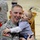 432nd CA BN prepares for deployment to Afghanistan A U.S. Army soldier in ACU (Army Combat Uniform) holds a young toddler, approximately 12-18 months old, who is leaning in close to the soldier's face. The soldier is smiling warmly, suggesting a reunion or family gathering event. The setting appears to be an indoor gymnasium or assembly hall.
