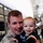 432nd CA BN prepares for deployment to Afghanistan A smiling male soldier in US Army ACU (Army Combat Uniform) digital camouflage is holding a young toddler, approximately 1-2 years old, who is also smiling at the camera. The setting appears to be an indoor military facility or hangar, with other people visible in the blurred background. The scene strongly suggests a homecoming or reunion event.