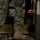 The image shows military personnel inside what appears to be a military transport aircraft or vehicle. One soldier in ACU (Army Combat Uniform) camouflage pattern is standing, visible from the waist down, while a second person's face is visible peering through a narrow opening or hatch. The scene appears to be inside a cargo hold with strapped-down equipment and wooden pallets.