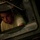 A young male with a short haircut is peering through a small rectangular window or hatch of what appears to be a military vehicle. The frame is constructed of riveted olive-drab metal, consistent with military equipment. The lighting is dim and dramatic, casting shadows across the scene.