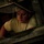 A young man in a light-colored military-style t-shirt is visible through a rectangular hatch or access panel of what appears to be a military vehicle or aircraft. He is leaning forward and looking downward, seemingly focused on a task. The image is shot from a low angle through the metal-framed opening, creating a dramatic, confined perspective.