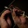 A close-up photograph of a pair of hands manipulating a thin white cord or rope through a metal carabiner or hook attached to a larger twisted wire cable. The image is taken in low-light conditions with dramatic chiaroscuro lighting emphasizing the hands and hardware. The activity appears to be rigging, securing, or tying a knot in a technical or industrial context.