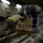 A heavily motion-blurred image showing what appears to be one or more individuals working with or moving cargo/boxes in an industrial or warehouse-like environment. The zoom-burst blur effect makes precise identification difficult, but a person wearing a light-colored shirt with a yellow high-visibility armband is visible leaning over a strapped cargo pallet or crate. The setting appears to be an aircraft cargo hold or loading facility based on the structural elements visible in the background.