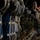 A military service member in ACU (Army Combat Uniform) camouflage pattern is working inside what appears to be a large military cargo aircraft, likely a C-17 or C-130. The individual is securing or checking static lines and rigging attached to multiple parachute bundles or cargo loads lined along the aircraft's interior wall. The scene appears to be a nighttime pre-jump or airdrop preparation operation.