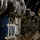 A military service member in ACU (Army Combat Uniform) camouflage pattern is working inside what appears to be a military cargo aircraft, securing or attaching static lines or rigging cables overhead. The aircraft cargo bay is loaded with multiple rows of military equipment bundles, parachute packs, and supply pallets secured with cargo straps. The scene appears to be a nighttime airdrop preparation operation.