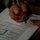 A person is filling out a structured form or document using a ballpoint pen, held on what appears to be a clipboard or binder. The image is taken at an angle, making the text on the form appear rotated. The lighting is dim with the subject's hands and the document being the primary focus.