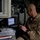 A bald male military personnel wearing a tan/khaki flight suit with a US flag patch on the right shoulder is seated at a workstation inside what appears to be a military aircraft. He is holding a small electronic device and reviewing documents, with a laptop computer open in front of him. The workstation is surrounded by various control panels and equipment typical of a military aircraft interior.
