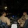 Two military aviators in tan/khaki flight suits are seated in the cockpit of a large military aircraft, likely a C-17 Globemaster III, engaged in conversation. The cockpit is illuminated by instrument panel lighting in a dark environment, suggesting nighttime operations. The pilot on the right displays a US flag patch on his right shoulder, indicating US Air Force personnel.