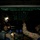 Two military aviators are seated in the cockpit of a large military aircraft during nighttime operations. The cockpit is illuminated primarily by green instrument lighting on the overhead panel, with blue-lit multifunction displays visible on the center console. One crew member is pointing at the overhead panel while the other observes, suggesting a checklist or systems review procedure.