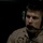 A young male military aviator or aircrew member is photographed in low-light conditions wearing a flight suit and aviation headset with boom microphone. He appears to be on duty, possibly aboard an aircraft, with his gaze directed slightly off-camera. The dramatic lighting highlights his face against a dark background, suggesting an operational or in-flight environment.