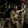 A military aircrew member in tan/khaki flight suit stands inside the cargo hold of a military transport aircraft, likely a C-17 or C-130. He is wearing green aviation headset with a boom microphone and reaching toward equipment or controls on the aircraft's interior wall. The cargo bay contains secured loads, tie-down straps, and parachute rigging lines.
