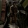 A military aircrew member in tan/desert flight suit stands inside the cargo hold of a military transport aircraft, flanked by large green cargo bundles or equipment packages. The individual is wearing a flight helmet with communications gear, a harness system, and tactical vest. The interior shows the aircraft's metal flooring, wall-mounted control panels, and tightly packed cargo on both sides.