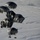 The image shows multiple large dark military cargo parachutes descending over a snow-covered landscape. Each parachute appears to be carrying military equipment or supply bundles suspended beneath them. The aerial perspective provides a clear view of the parachute deployment pattern against the white terrain below.