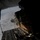 The image is taken from inside a military cargo aircraft with its rear ramp open during flight. A cargo pallet or supply bundle is in the process of being airdropped, with parachute deployment lines visible. The terrain below appears to be snow-covered or cloud-covered, suggesting a cold-weather or high-altitude operation.