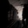 The image shows the interior of a military cargo aircraft with the rear ramp open, captured in high-contrast silhouette lighting. A crew member in flight gear stands near the open ramp while a large cargo pallet with netting appears to be in the process of being airdropped. Bright exterior light floods through the open ramp, creating strong silhouette effects against the dark interior.
