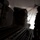The image shows the interior of a military cargo aircraft with its rear ramp open during what appears to be an aerial delivery or airdrop operation. A silhouetted crew member stands near the open ramp while a large wrapped cargo pallet is in the process of being deployed. The bright exterior light contrasts sharply with the dark aircraft interior, creating a high-contrast silhouette effect.