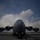 A large military transport aircraft, consistent with a Boeing C-17 Globemaster III, is photographed from a dramatic low front-facing angle on a tarmac. The image is taken in low-light or overcast conditions, rendering the aircraft in deep shadow against a partly cloudy sky. The wide-angle perspective emphasizes the aircraft's four turbofan engines and massive fuselage.