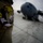 A close-up photograph of a military uniform sleeve bearing a patch reading '816 EAS' (Expeditionary Airlift Squadron), featuring a skull and sun/halo design. In the blurred background, a large military transport aircraft — consistent with a C-17 Globemaster III — is visible on a flight line or tarmac. The image uses shallow depth of field to emphasize the unit patch.