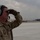 A military airman wearing a tan flight suit and green hearing protection headset stands on an airfield tarmac, shielding his eyes with a gloved hand while looking into the distance. He displays unit patches including one labeled '876 EAS' on his left arm and additional patches on his chest. In the background, a large military transport aircraft (likely a C-17 Globemaster) is visible on the runway under an overcast sky.