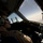 A military pilot is seated in the cockpit of an aircraft in flight, wearing a tan/khaki flight suit, aviation headset, and sunglasses. The cockpit windows reveal a bright sky with scattered clouds, suggesting high-altitude flight. The pilot appears to be monitoring instruments or scanning outside the aircraft.