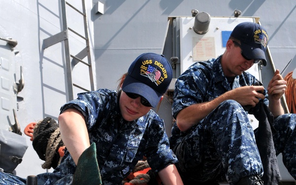Two U.S. Navy sailors in NWU (Navy Working Uniform) digital camouflage are performing maintenance or cleaning on what appears to be a machine gun (likely an M2 .50 caliber or M240) on the deck of a naval vessel. Both sailors are wearing USS Decatur (DDG-73) ship's ball caps. The work is taking place on an open deck with ship superstructure visible in the background.
