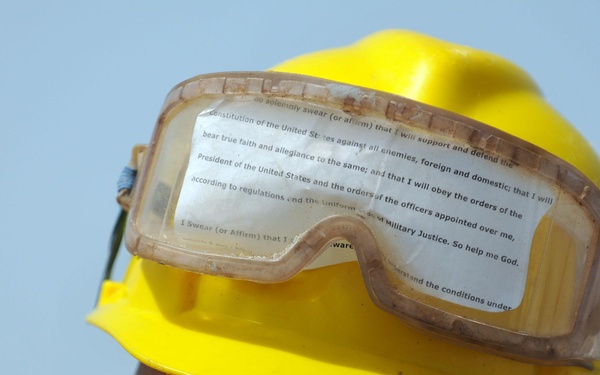 A person wearing a yellow hard hat with safety goggles resting on top and Ray-Ban sunglasses. A printed text is affixed inside the goggles lens, visible as the U.S. Military Oath of Enlistment. The image is a close-up portrait shot against a light blue sky background.