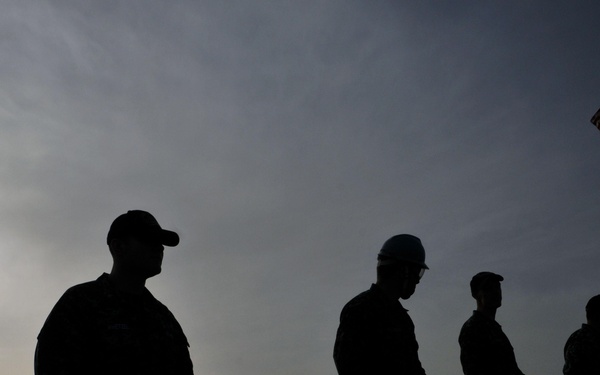 A silhouetted group of five military personnel stand in a line on the deck of a naval vessel, holding a line or rope. An American flag is visible flying from a mast in the background on the right side. The image is taken at dawn or dusk with a hazy sun visible on the left horizon, with a distant cityscape or port visible in the background.