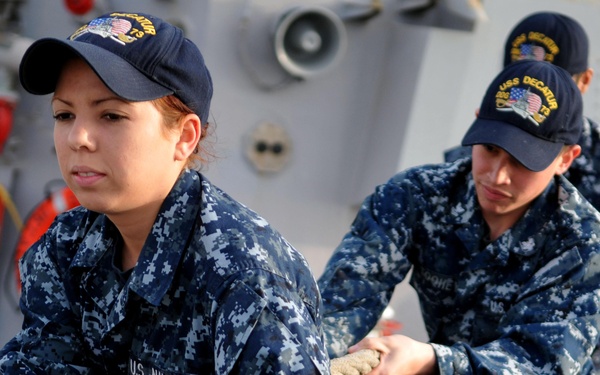 Two U.S. Navy sailors in Navy Working Uniforms (NWU) are hauling a mooring line on the deck of USS Decatur, as indicated by the ship name visible on their caps. The sailors are engaged in line-handling operations, likely during a mooring or unmooring evolution. The image is taken on the ship's weather deck with naval equipment and safety gear visible in the background.